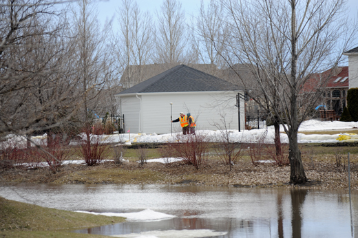 Flooded ground in the front, in the distance two survey workers check the height of white sand bags in front of a neighborhood.