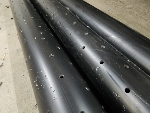 Black plastic pipes with holes cut out for water drainage every few inches sitting on cement waiting to be installed.