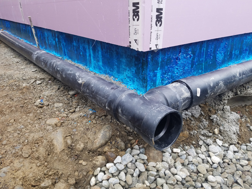 Black plastic pipes installed against the base of a home waiting to be backfilled with gravel and topsoil.