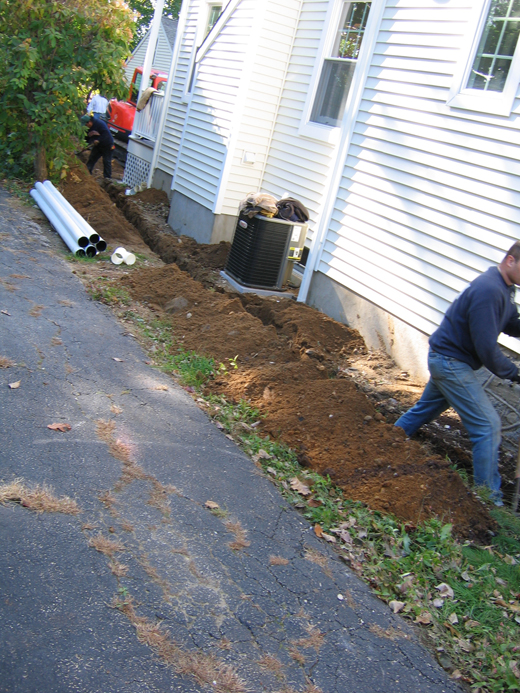 Trench dug around a house with a pipe sitting to the side waiting to be installed in the trench.