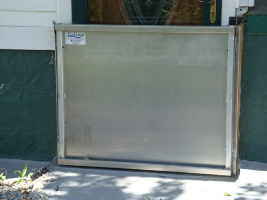 A stainless steel opening barrier closure to block floodwater entry from a wooden resdiential front door with decorative glass.