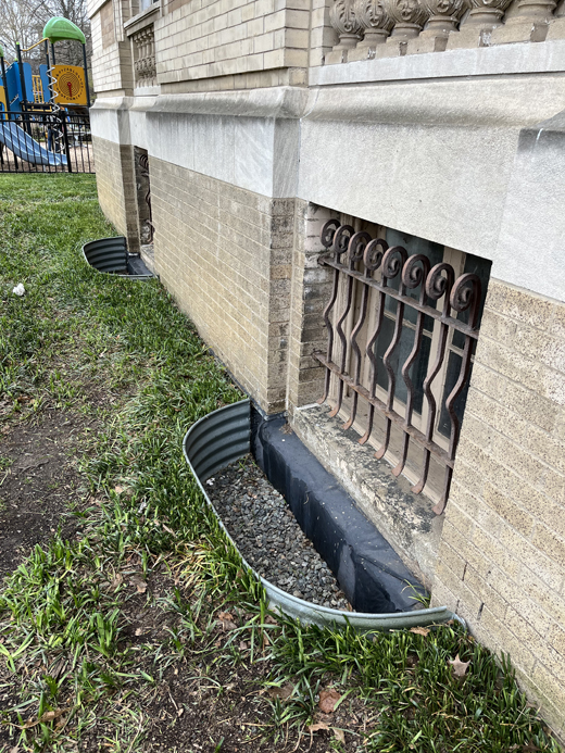 Metal floodwall beneath a basement window covered with decorative metal window bars, with waterproof membrane also visible.