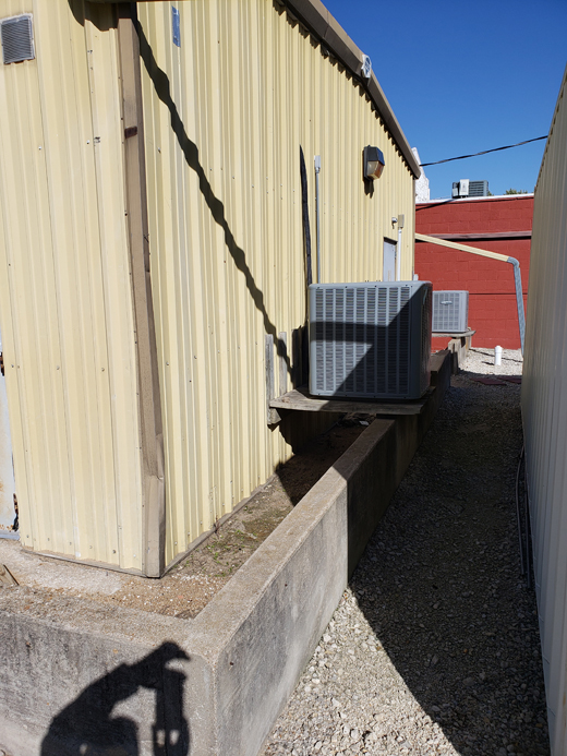 To protect against shallow flooding, a floodwall surrounds a yellow metal building, with elevated utilities in the rear. The cement wall has grooves in it at the openings for drop in barriers.