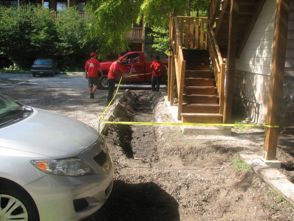 Three workers preparing a site along the edge of a parking lot for a bioswale, caution tape marks the edge of the site