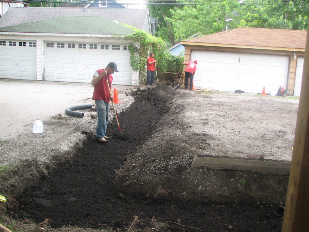 Three workers using shovels to prepare soil for a bioswale between two garages/driveways