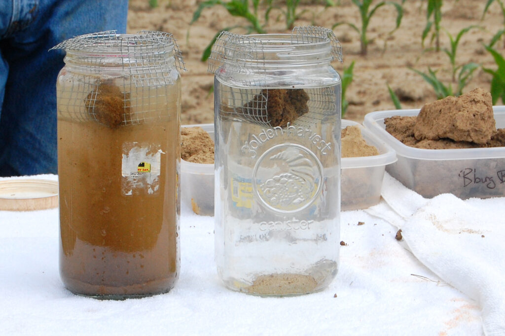 Two glass jugs on a table the left of which shows the impact of soil under conventional tillage on water (the jug is full and murky brown) and the right of which shows the impact of soil under no-till agriculture (the jug is effectively empty)