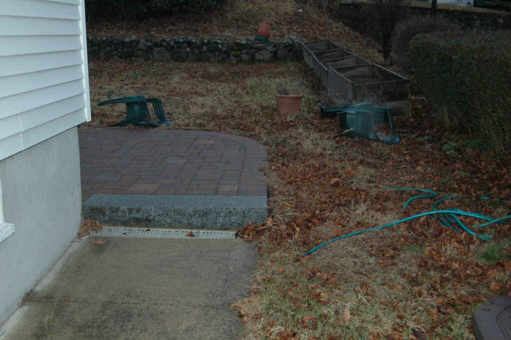 A side yard where a drywell has been installed, beyond is a backyard covered in brown fall leaves.
