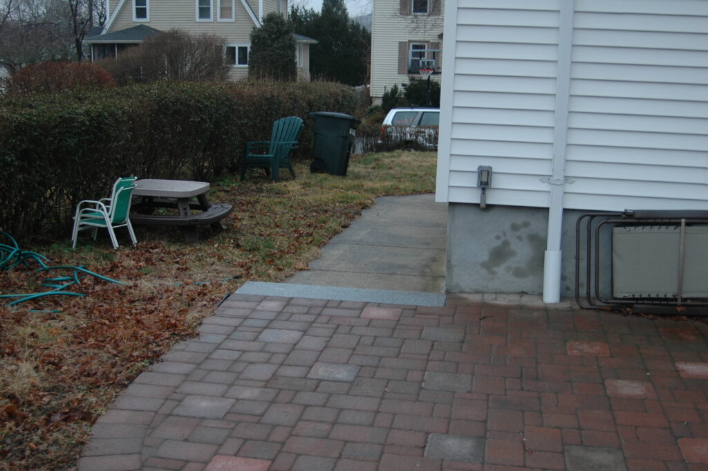 A side yard where a drywell has been installed, beyond is the street. The side yard has a square, plastic picnic table, chairs, and a trash bin.