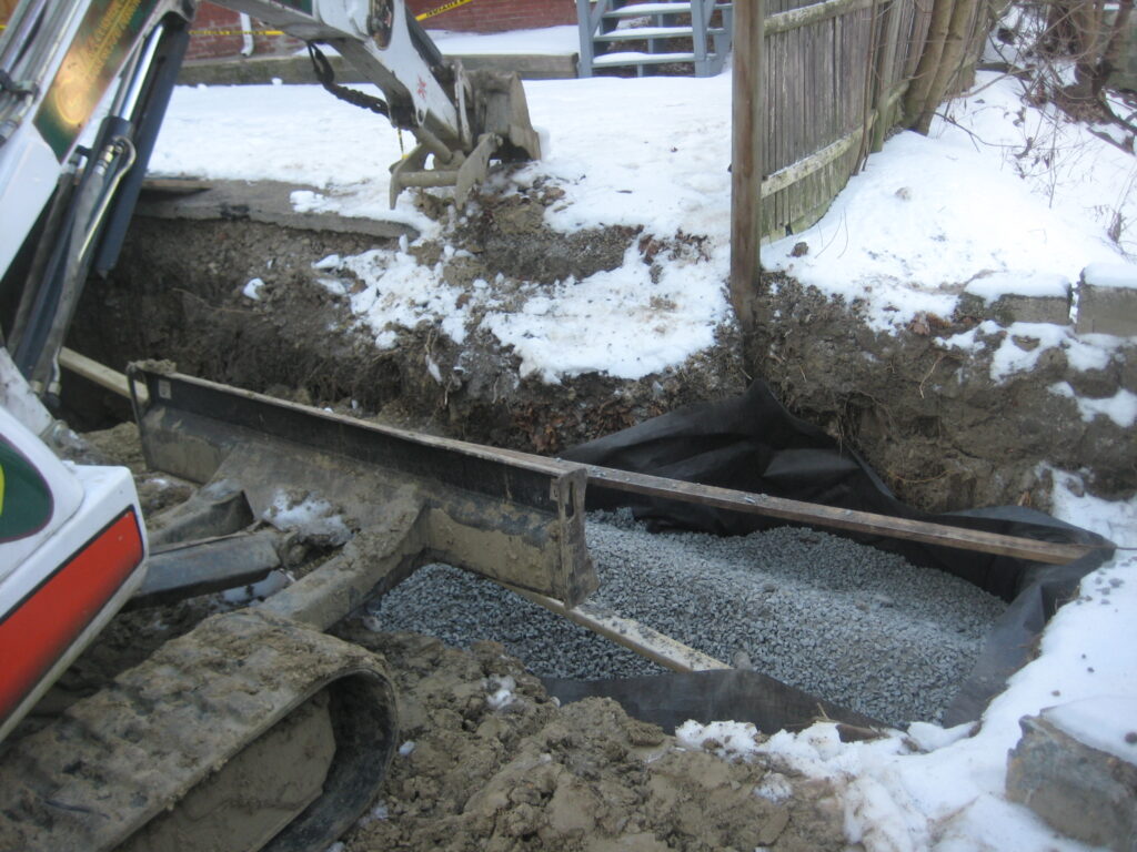 A construction vehicle is resting alongside a long and wide rectangular drywell hole that is filled with pea gravel. The hole is around six feet deep below the driveway.