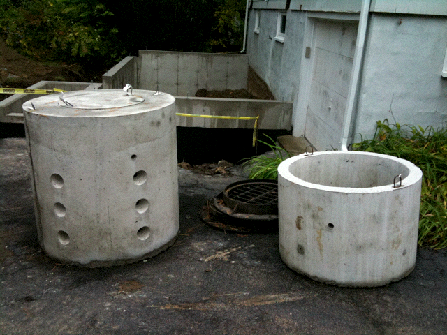 Two pieces of the dry well sitting on the ground next to a building waiting to be installed.