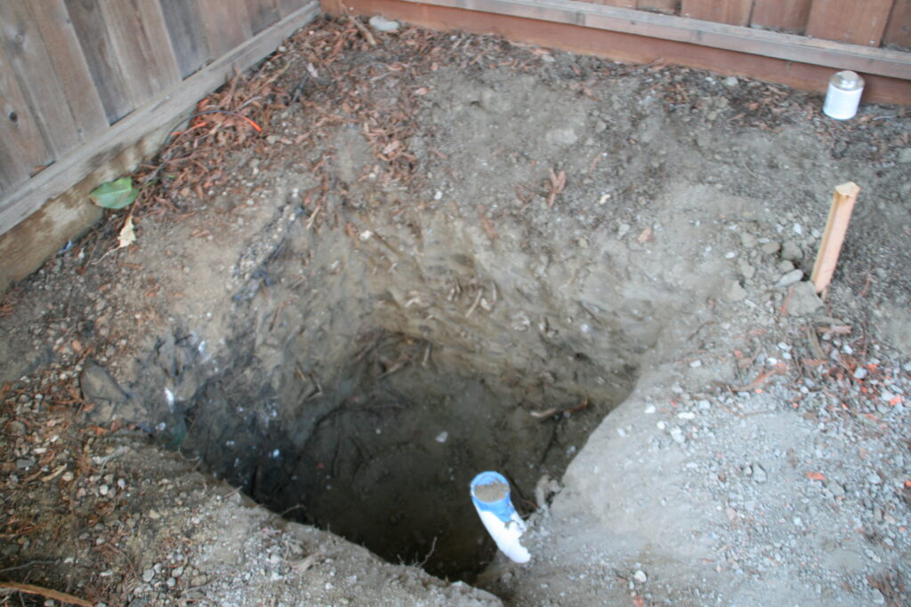 A square hole in the ground likely around 2-foot by 2-foot with a pipe coming out of the side.