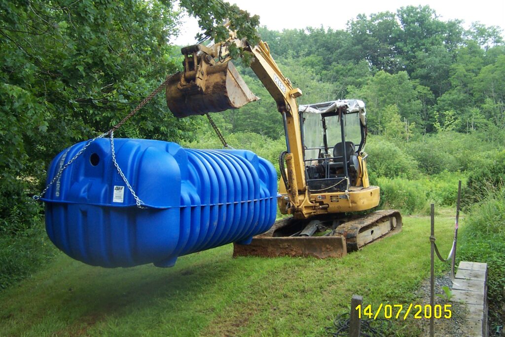 A large blue septic tank is raised off the ground by a construction vehicle in preparation for installation.
